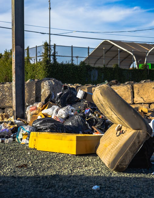 A large green wheeled waste container filled with several red and white bags of building or construction materials, positioned outdoors on a paved surface against a dark brick wall. The container appe
