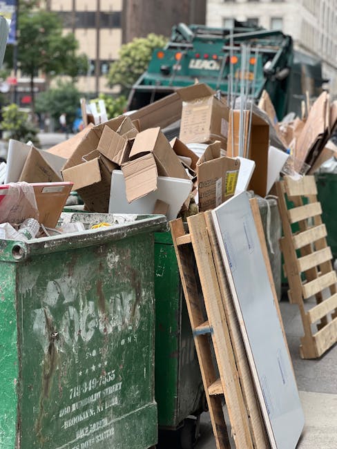 An outdoor scene showing a large green rubbish bin overflowing with various types of waste, including multiple cardboard boxes, some flattened and others still in three-dimensional form, alongside sma