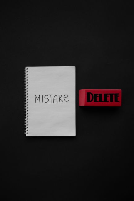 A black background with a white spiral-bound notebook positioned towards the left, displaying the handwritten word 'MISTAKE' on its cover. To the right of the notebook, there is a red rubber stamp wit