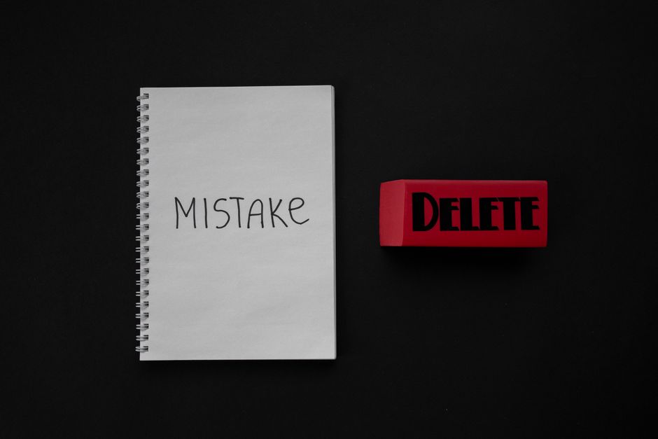 A black background with a white spiral-bound notebook positioned towards the left, displaying the handwritten word 'MISTAKE' on its cover. To the right of the notebook, there is a red rubber stamp wit