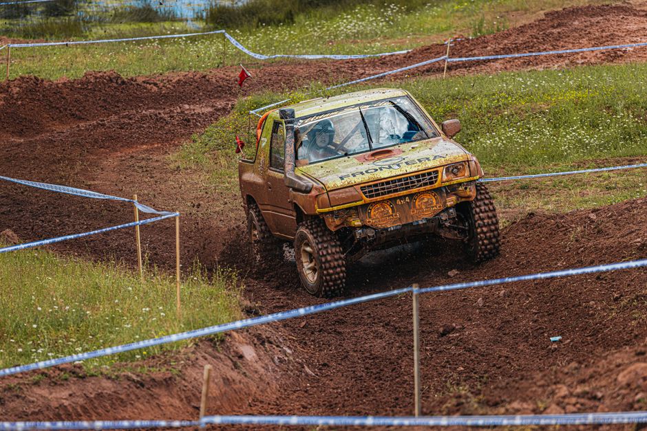 A rugged grey Toyota off-road vehicle with a black roof rack and additional lighting attached, navigating through a muddy forest trail surrounded by dense green foliage and trees. The vehicle's front 